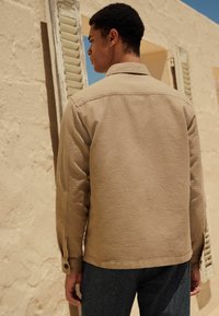Beige strukturierte Jacke mit Kragen und geknöpften Manschetten, kombiniert mit dunkler Jeans. Der Hintergrund zeigt hellfarbene Wände und Fensterläden.