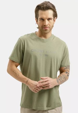 Mężczyzna o krótkich brązowych włosach, noszący jasnozielony t-shirt "Cross Jeans", pokazujący tatuaże na obu rękach, stojący ze złączonymi dłońmi.