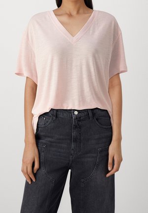 Kobieta w jasnoróżowym t-shircie z krótkim rękawem i dekoltem w serek, wsuniętym w czarne, wysokie jeansy, pokazana od ramion do bioder.