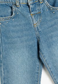 Jeans de denim azul claro con una textura deslavada, que presentan tachuelas plateadas en los bolsillos y costuras en contraste de color naranja.