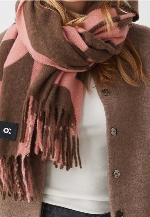 Femme portant une grande écharpe à motifs roses et marron avec des franges, par-dessus un cardigan marron boutonné et un haut blanc.