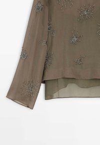 Gennemsigtig brun bluse med lange ærmer, prydet med sølvperlede edderkopmønstre. Har et lagdelt design med en let gennemsigtig ryg.