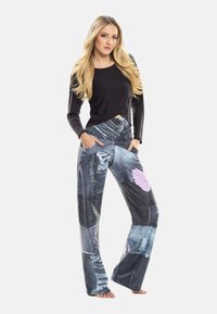 Sieviete ar gariem blondiem matiem valkā melnu garroku krekliņu un plašu bikses ar zilas džinsa plankuma apdruku un rozā sirds detaļu, stāv basām kājām.