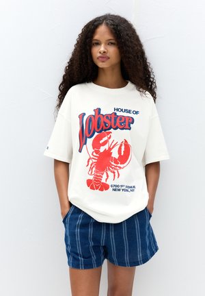 T-shirt blanche avec un graphique de homard rouge et du texte bleu, associée à un short rayé bleu marine. Coupe décontractée avec des manches larges et un col rond.