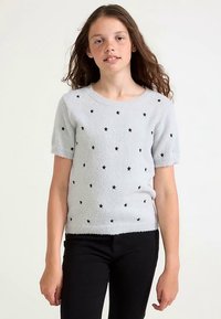 Maglione grigio chiaro a maniche corte con una texture pelosa e motivi a stelle nere. Indossato con pantaloni neri.