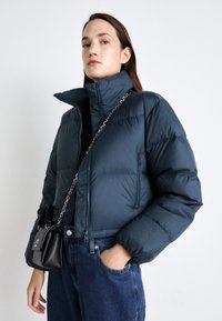 Marineblå pufferjakke med høy krage, glidelås og frontlomme; brukt med en svart crossover-veske og blå denim. Glatt tekstur.
