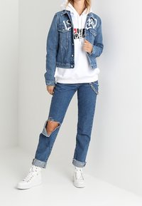 Giacca di jeans con scritte bianche, abbinata a una felpa bianca con cappuccio e jeans strappati con risvolto. Sneakers bianche alte completano l'outfit.