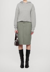 Persona con sudadera gris, falda verde oliva hasta la rodilla con bolsillos frontales, botas negras hasta la rodilla y sosteniendo un pequeño bolso oscuro.