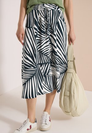 Kvinde iført sort og hvid abstrakt stribet midinederdel, hvide sneakers, der holder en stor beige skuldertaske.