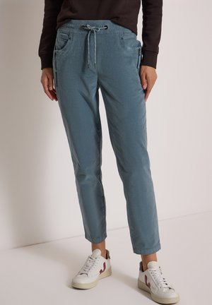 Personne portant un pantalon bleu à cordon avec poches avant et des baskets blanches avec des accents roses, debout devant un fond neutre.