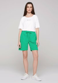 Camicia bianca a maniche corte con logo stampato e pantaloni corti verdi con cintura elastica e tasche, abbinati a sneaker bianche.