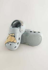 Light gray clogs with fuzzy lining, featuring a cartoon croissant and "BONJOUR" text on the front, a textured sole, and ventilation holes.