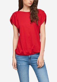 T-shirt rossa in cotone con maniche corte a sbuffo e orlo arricciato, abbinata a jeans blu strappati. Texture morbida e vestibilità rilassata.