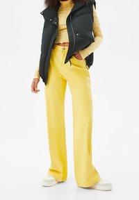 Yellow wide-leg denim pants with a smooth texture, paired with a black puffer vest and light colored top, set against a white background.