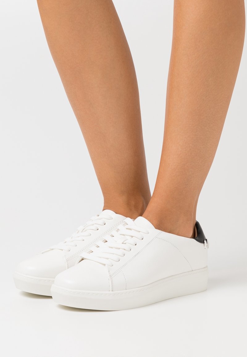 Baskets basses blanches avec lacets et détail noir au talon portées pieds nus sur un fond uni.