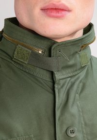 Guler din țesătură verde cu fermoar și închidere cu velcro. Prezintă un detaliu cu un buton dedesubt, arătând o finisare netedă, texturată.