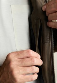 Braune Lederjacke mit Reißverschluss vorne, Innentasche und strukturierter Oberfläche, getragen über einem hellen Hemd. Hand greift die Jacke.