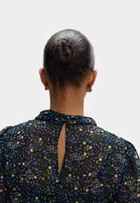 Czarna blouse w kwiaty z niebieskimi, żółtymi i zielonymi wzorami. Detal w kształcie dziurki z tyłu, zgromadzony dekolt i gładka, miękka faktura. Włosy upięte w kok.