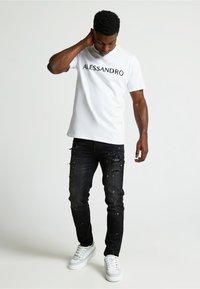 Camiseta blanca de algodón con "ALLESSANDRO" impreso en negro, combinada con jeans negros desgastados y zapatillas blancas. Estilo casual de streetwear.