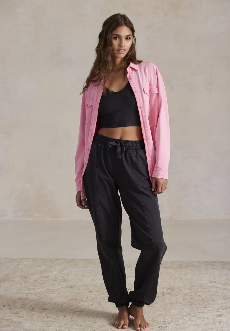 Camicia button-up rosa chiaro sopra un crop top nero, abbinata a jogger neri con cordino. Tessuto morbido, vestibilità rilassata e caviglie a maniche.