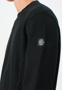 Mężczyzna w czarnym swetrze o prążkowanej strukturze, z małą okrągłą naszywką logo Guess na górnej części rękawa na jednolitym tle.