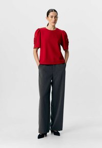 Haut rouge à manches courtes avec des manches bouffantes et des accents décoratifs. Associé à un pantalon gris taille haute à jambes larges et des chaussures à talons noirs.