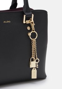 Zwarte handtas met textuur, gouden hardware, kettingaccenten en een charm met een hangslot en sleutel. Merklogo is zichtbaar.