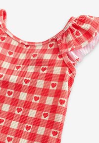 Tessuto a quadretti rosso e bianco con texture in rilievo e piccoli motivi a cuore. Presenta dettagli arricciati sulle spalle e un collo rotondo.