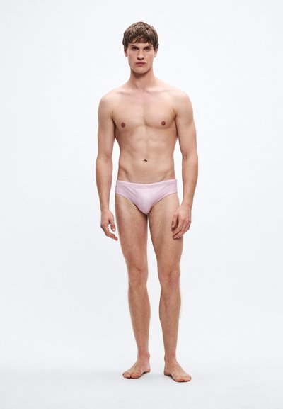 Hele roosa ujumispüksid, millel on sile tekstuur ja minimalistlik disain, madal lõige ja elastne vöökoht. Modell seisab paljajalu.