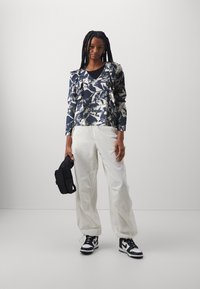 Top navale e bianco con motivo floreale e ruches; abbinato a pantaloni bianchi larghi e sneaker nere; tiene in mano una piccola borsa nera.