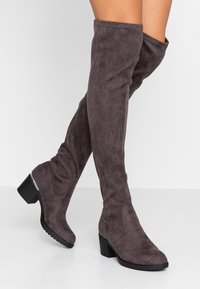 Botas por encima de la rodilla en ante gris suave con tacón ancho y suela de goma negra texturizada. Presentan detalles cosidos en la caña.