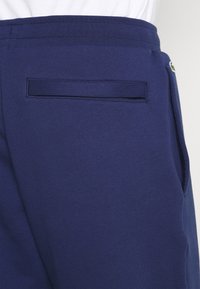 Gros plan sur un pantalon bleu avec une poche arrière passepoilée et une poche latérale ornée d’un petit logo vert à la ceinture.