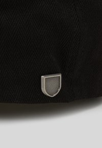 Casquette noire avec un motif en chevrons et un petit emblème en argent, comportant un design de lignes diagonales texturées au centre.