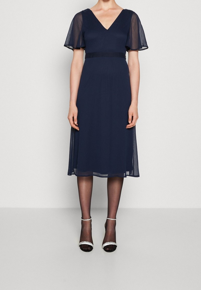 Marinblå A-linje klänning med V-hals, genomskinliga fladderärmar och ett sidentyg midjeband. Matchas med svarta klackar med remmar och genomskinliga strumpbyxor.