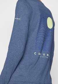 Modrá dlouhá košile Columbia Sportswear s grafickým designem žlutého kruhu a vlnitých modrých linií na zadní straně, a logem Columbia na rukávu.