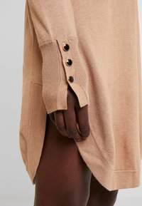 Maglione lavorato a maglia color marrone chiaro con maniche a costine, caratterizzato da bottoni color oro sui polsini e fessure laterali per una maggiore libertà di movimento.