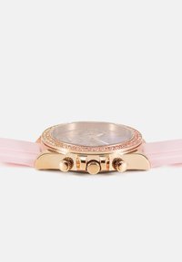 Montre chronographe ronde avec un boîtier en or rose, une lunette ornée de pierres roses, et un bracelet en silicone doux rose. Comprend trois boutons de couronne sur le côté.