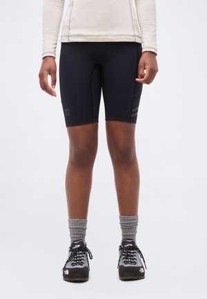 Personne portant un short de sport noir mi-cuisses, des chaussettes grises et des chaussures de randonnée noires et grises sur un fond uni.