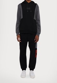 Hoodie i svart och grått med en logotyp på bröstet, parat med svarta sweatpants med en orange logotypdetalj och muddar vid anklarna.