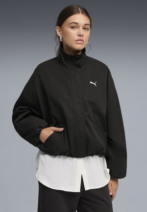 Giovane donna con capelli scuri indossa una giacca nera oversize Puma sopra una camicia bianca, in piedi con una mano in tasca davanti a uno sfondo grigio.