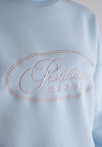 Sudadera azul claro con escote redondeado y logo bordado en rosa que dice "Prohibido" con estrellas, con una textura de tela suave.