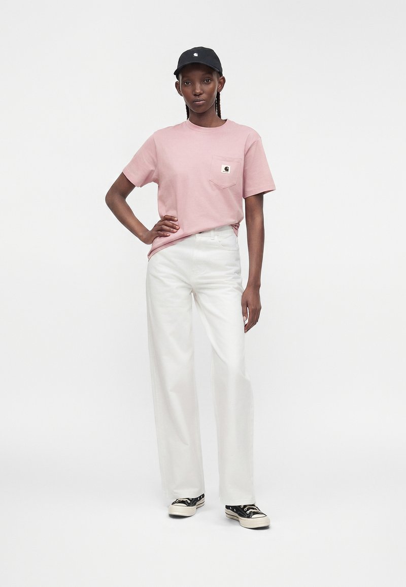 Young woman standing with hand on hip wearing pink T-shirt, white wide-leg pants, black cap, and black sneakers with white soles.