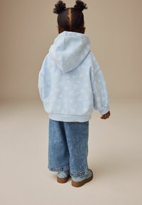 Lyseblå hoodie med subtilt blomstermønster, parret med brede denimjeans og blå blomsterstøvler på en neutral baggrund.