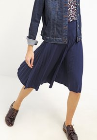 Marinblå veckad kjol med en flödande design, parat med en jeansjacka. Jackan har metallknappar och uppkavlade ärmar.