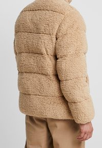 Tan, pluizige fleece pufferjack met horizontale stiksels en een relaxte pasvorm. Hoge kraag, geen zichtbare hardware, gestructureerd oppervlak.