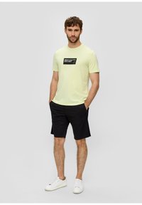 Light yellow cotton t-shirt with a black rectangular graphic on the chest, paired with dark shorts and white sneakers.