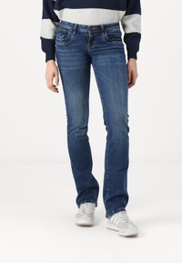 Vaqueros de denim azul con un corte recto, que cuentan con bolsillos frontales y un ligero desgaste. Combinados con zapatillas blancas y grises.