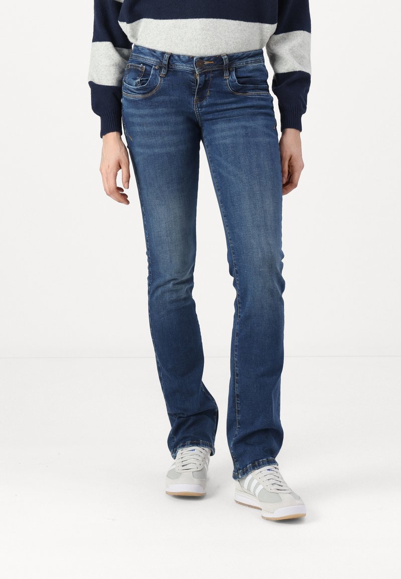 Vaqueros de denim azul con un corte recto, que cuentan con bolsillos frontales y un ligero desgaste. Combinados con zapatillas blancas y grises.