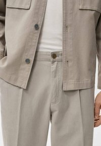 Veste légère beige avec deux poches avant, fermeture à boutons, et pantalon plissé beige assorti avec des boutons contrastants.