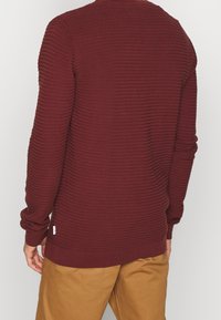 Bordeauxrode gebreide trui met verhoogde horizontale strepen, geribbelde boorden en zoom. Voorzien van een ronde halslijn en een zijlabel met merknaam.